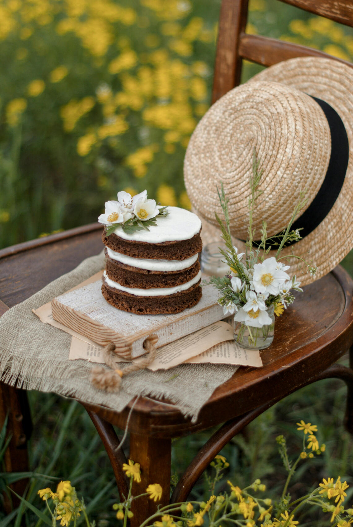 Small layered cake and straw hat on a brown chair surrounded by yellow flowers